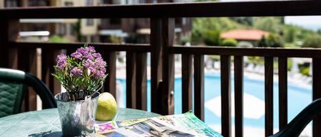 A table with a magazine, a potted plant, and a lemon on a balcony overlooking a swimming pool.
