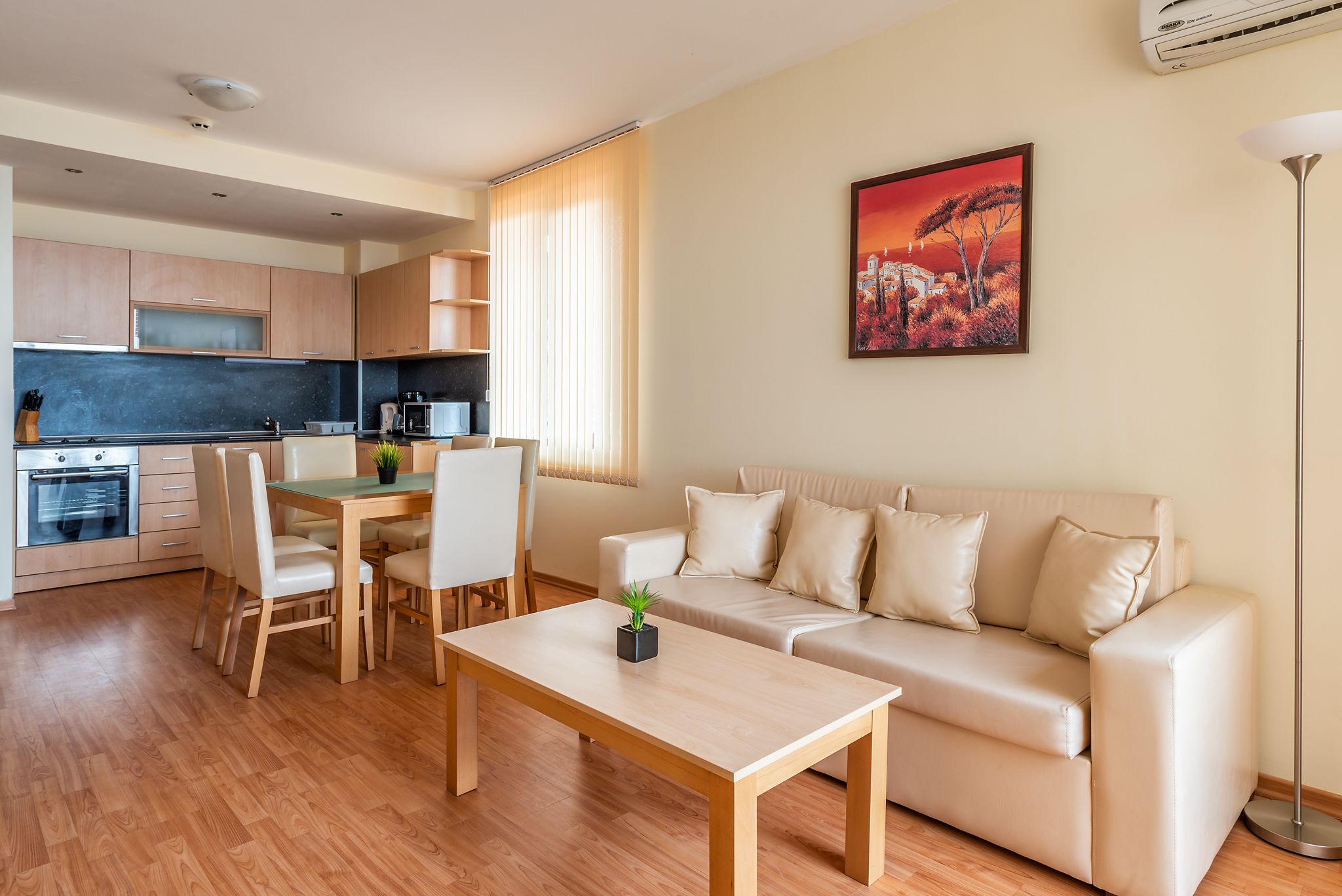 A modern living room with a beige sofa, a wooden coffee table, and a dining table with chairs. There is a kitchen area with cabinets and appliances in the background.