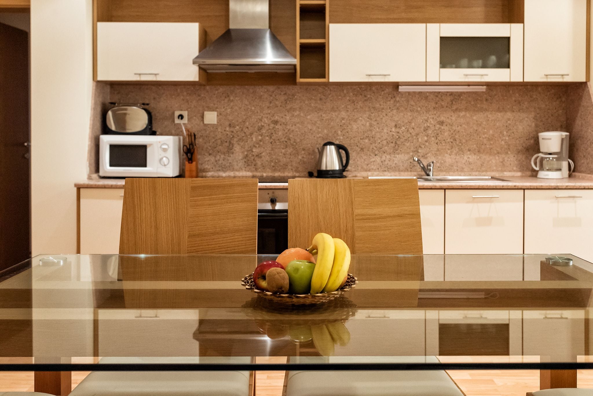 A modern kitchen with a glass dining table, wooden chairs, and a fruit bowl.