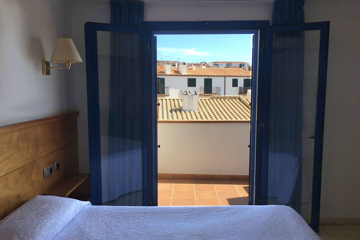 A hotel room with a bed, a nightstand, a lamp, and a view of the city through a door.