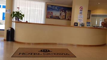 Zona de recepción de hotel con mostrador de recepción, área de registro, un ventanal con persianas, un cuadro enmarcado en la pared y un tapete de bienvenida con el nombre del hotel.