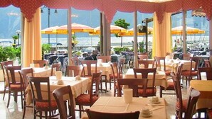 A restaurant with tables and chairs arranged for dining, large windows with view of the sea, and outdoor umbrellas. - Hotel Sole - Limone (Limone sul Garda)
