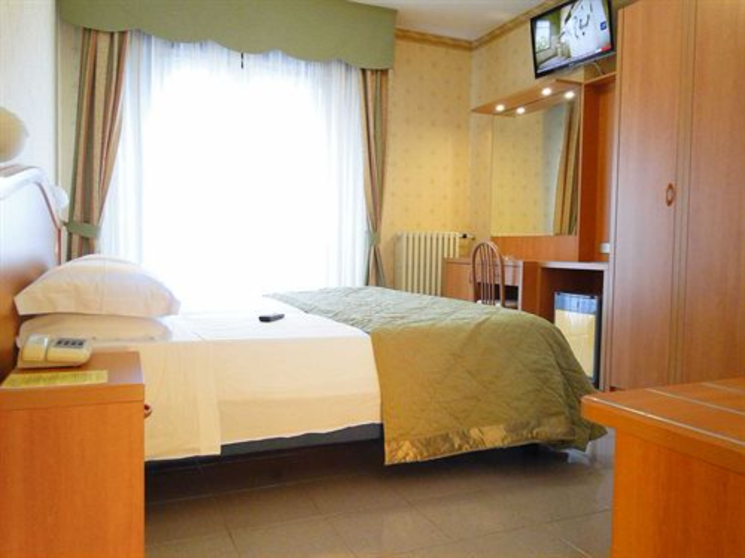 Una habitación de hotel con una cama, una mesita de noche con lámpara, un televisor montado en la pared y una ventana con cortinas.