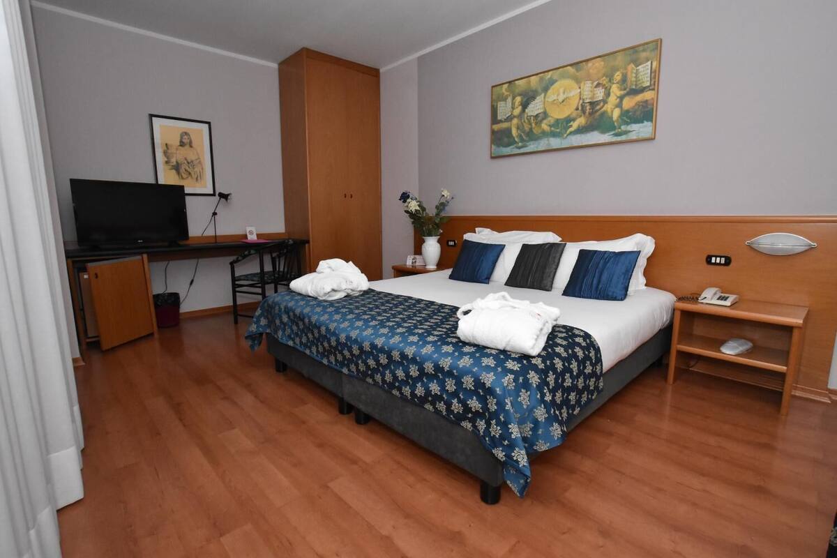 A hotel room with a bed, a desk with a TV, a wooden wardrobe, and a painting on the wall.