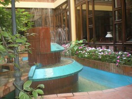 Una fuente con chorros de agua en un patio con piso de ladrillo y parterres de flores.