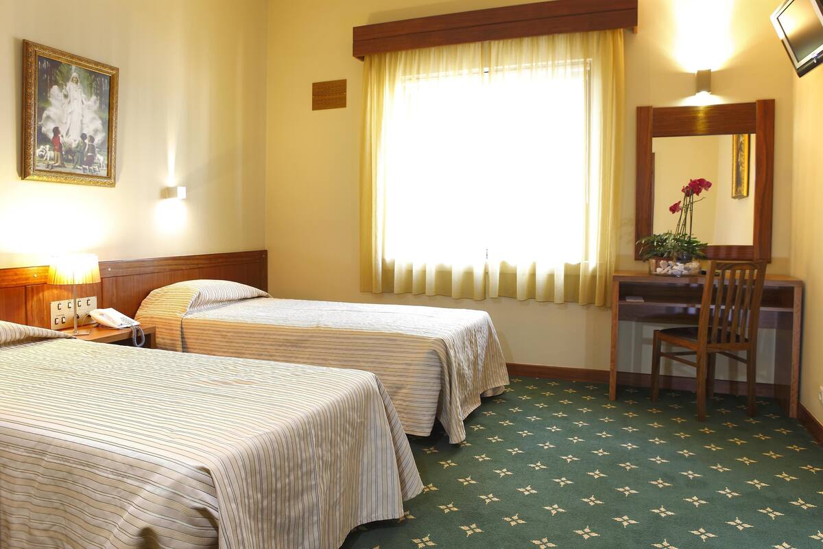 Habitación de hotel con dos camas individuales, un escritorio con silla, un espejo, un teléfono y una ventana con cortinas.