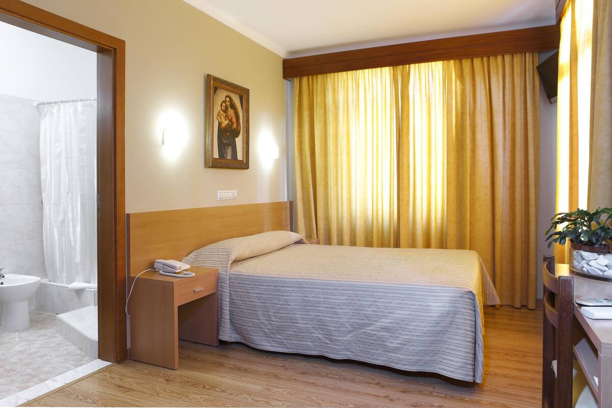 Habitación de hotel con cama, mesita de noche con teléfono, pequeño escritorio con planta y baño con cortina de ducha.