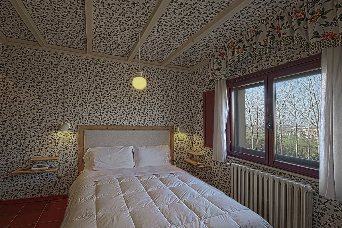A bedroom with floral wallpaper, a wooden bed, bedside lamps, a window with curtains, and a radiator.