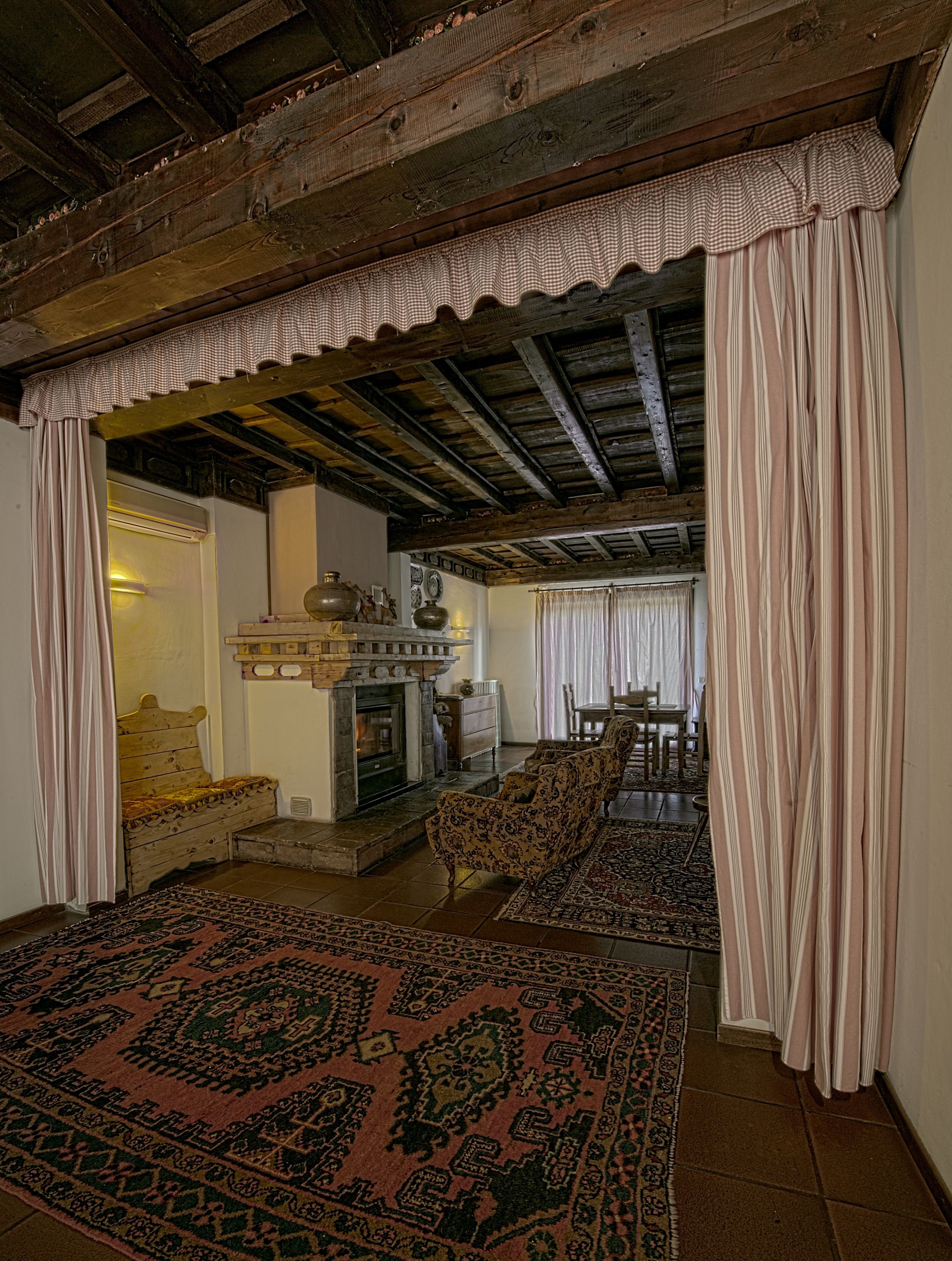 Quarto tradicional com lareira, vigas de madeira, tapete estampado e cortina xadrez.