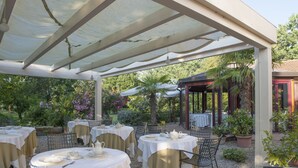 An outdoor patio with round tables set for dining, covered by a white canopy, surrounded by lush greenery and potted plants. - Villa Belfiore (Ostellato)