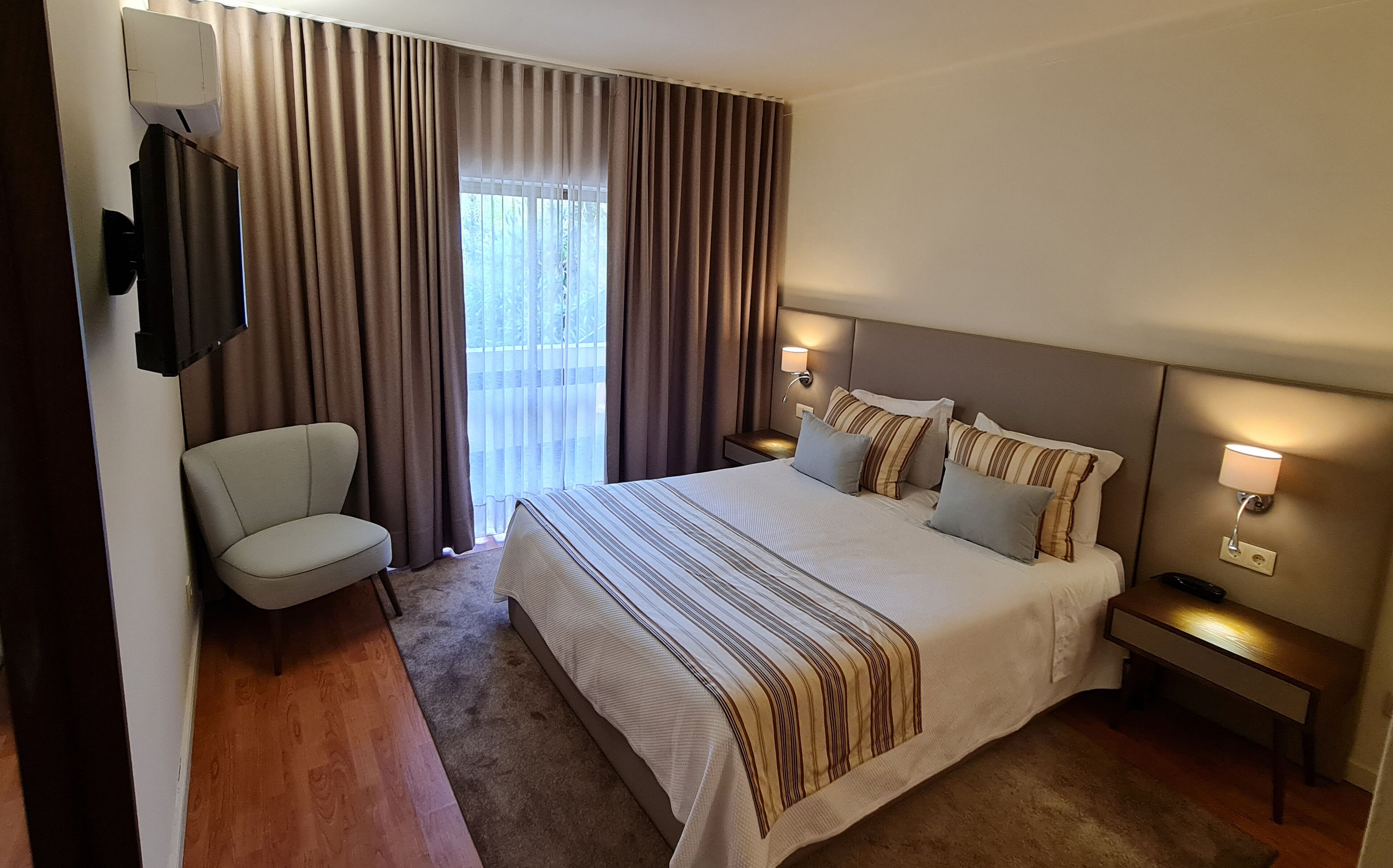 A hotel room with a large bed, two bedside tables with lamps, a chair, and a TV mounted on the wall.