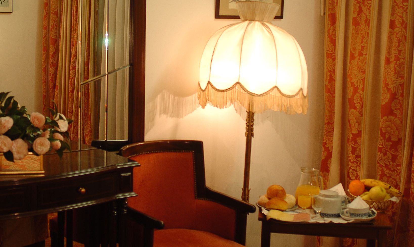 A room with a red upholstered chair, a table with fruits and a teapot, and a lamp with a white shade.