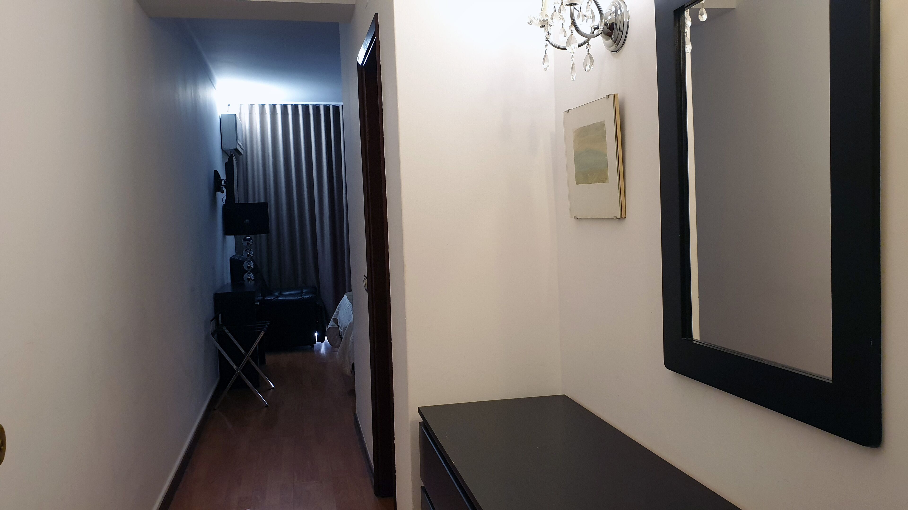 A narrow hallway with a wooden floor, a black desk with a lamp, a chair, and a small table. There is a door on the left and a mirror on the right.