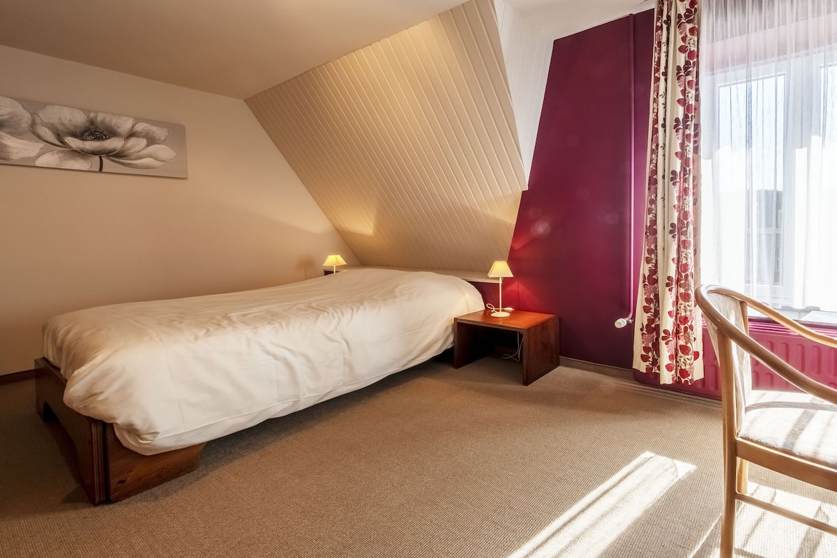 A bedroom with a single bed, a nightstand with a lamp, a window with sheer curtains, and a radiator.