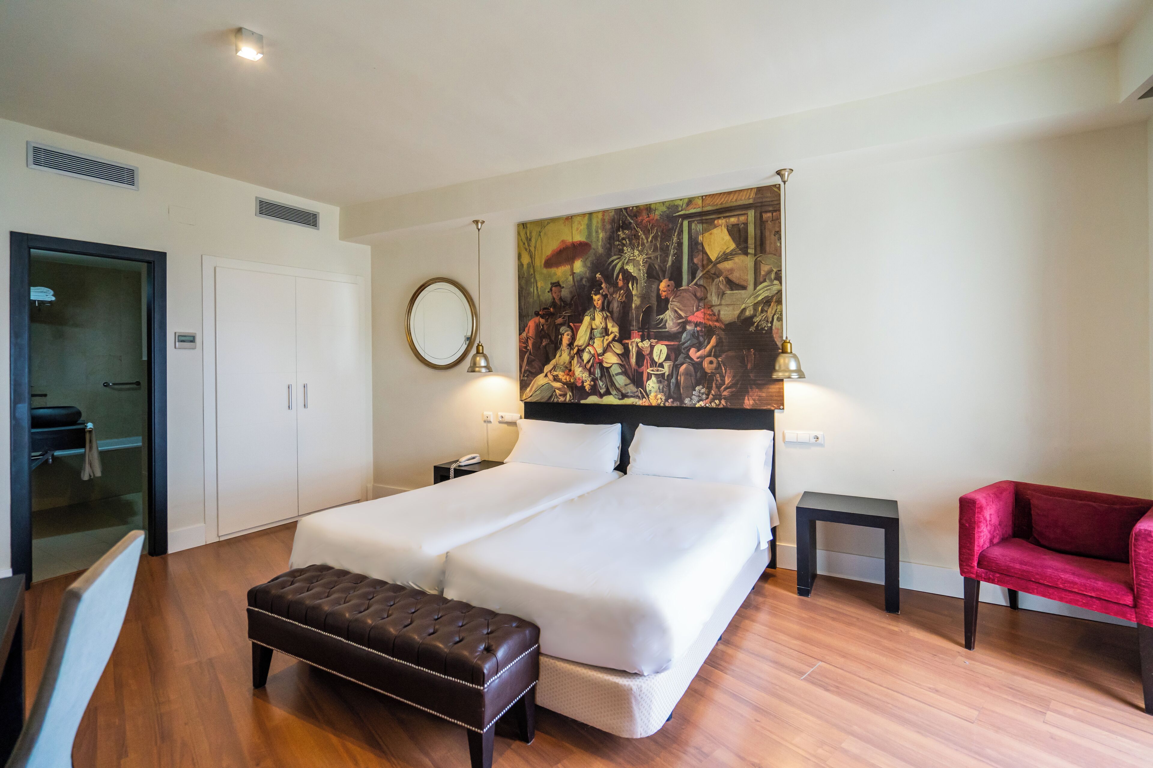 A hotel room with a large bed, a red chair, a painting on the wall, and a small table.