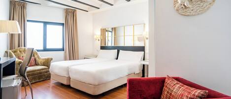 Habitación de hotel con cama doble, escritorio con lámpara, sillón rojo y ventana grande con cortinas.