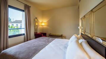 Habitación de hotel con cama, mesita de noche, lámpara, ventana con cortinas y vista a edificios.