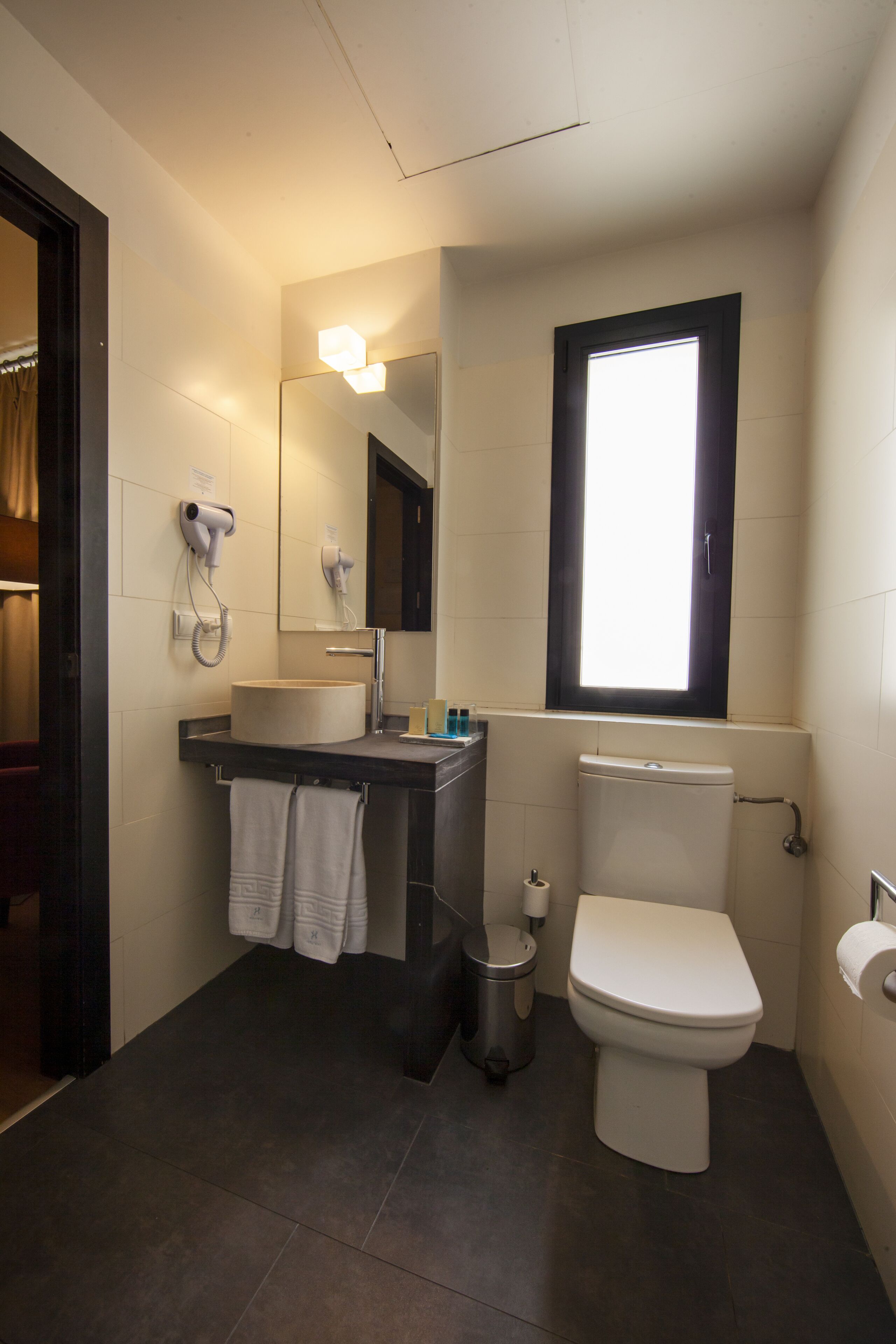 A bathroom with a toilet, sink, and mirror. There is a window above the toilet and a hairdryer on the wall.