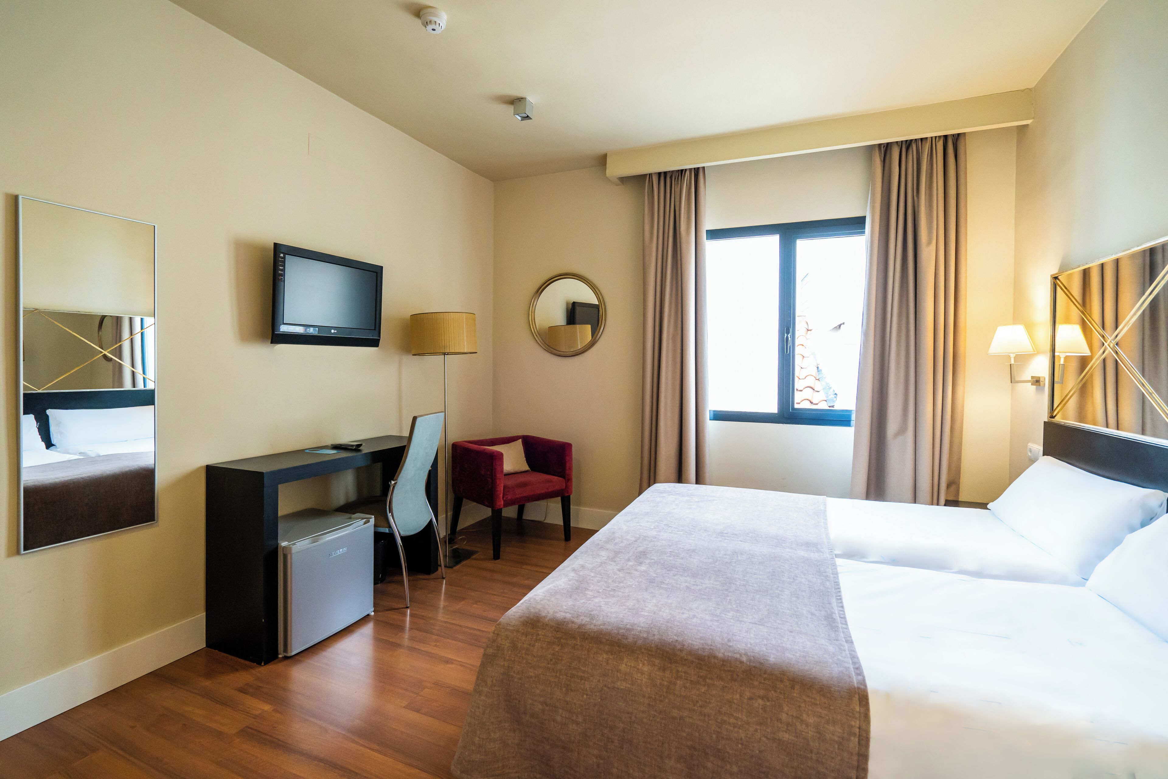 Habitació d'hotel amb un llit, escriptori amb ordinador, cadira, nevera petita, televisió i finestra amb cortines.