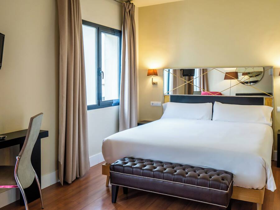 Habitación de hotel con una cama grande, un escritorio con silla, televisor y ventana con cortinas.
