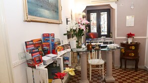 Una stanza con pavimento a scacchi, un tavolo in vetro, una sedia bianca, un vaso con fiori ed uno scaffale con opuscoli turistici.