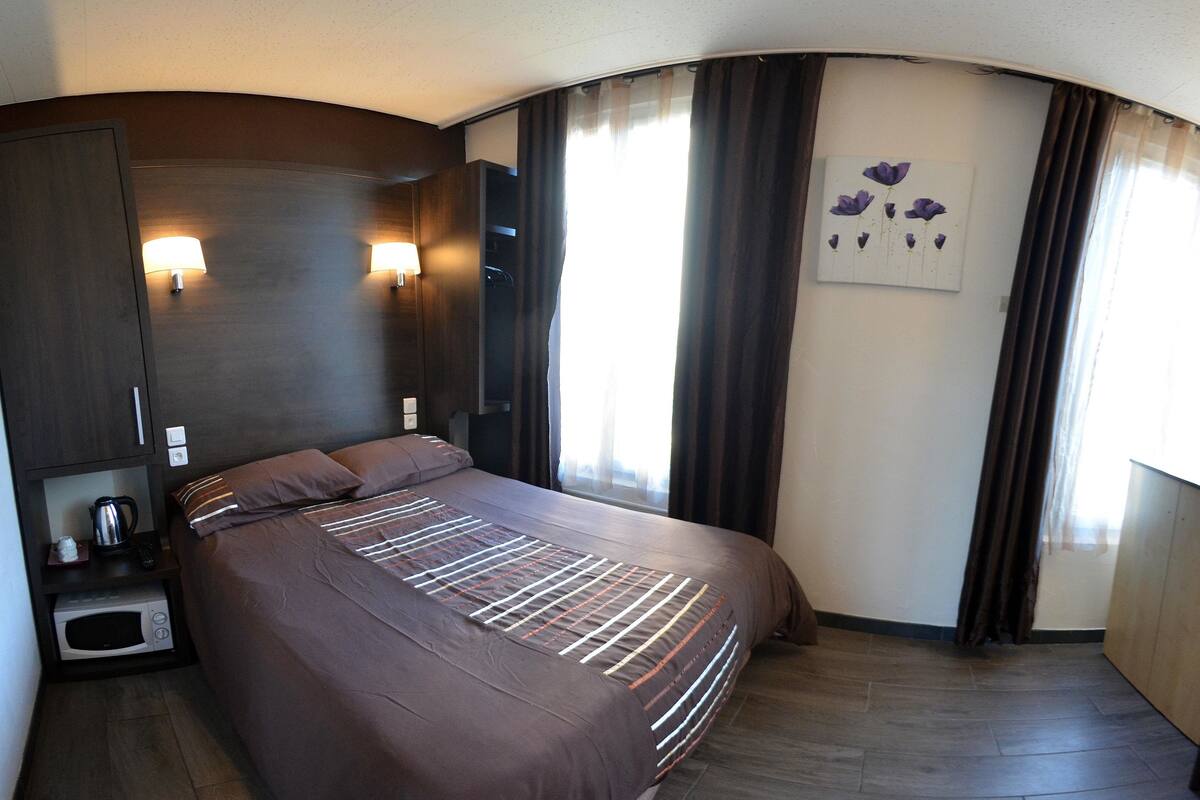 Habitación de hotel con una cama grande, una mesita de noche con hervidor y mando a distancia, un televisor de pared y una ventana con cortinas.
