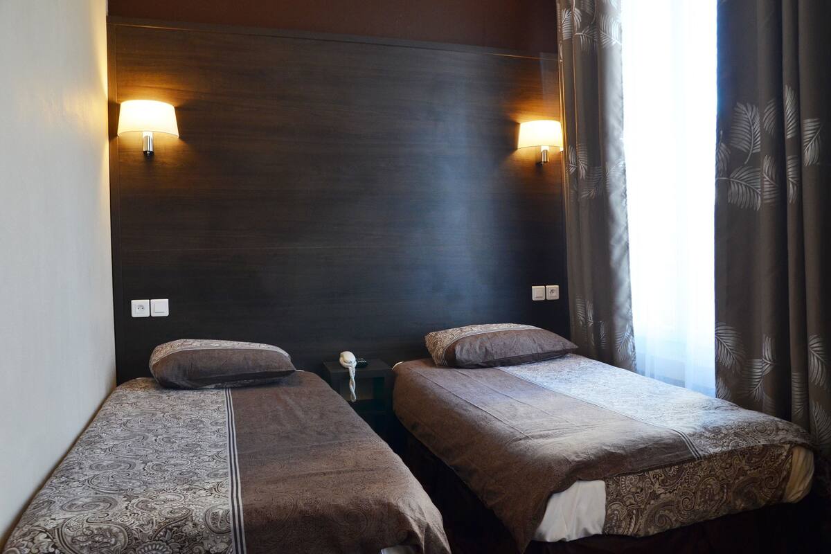 Habitación de hotel con dos camas individuales, cabecero oscuro, lámparas de pared, mesita de noche con lámpara y ventana con cortinas.