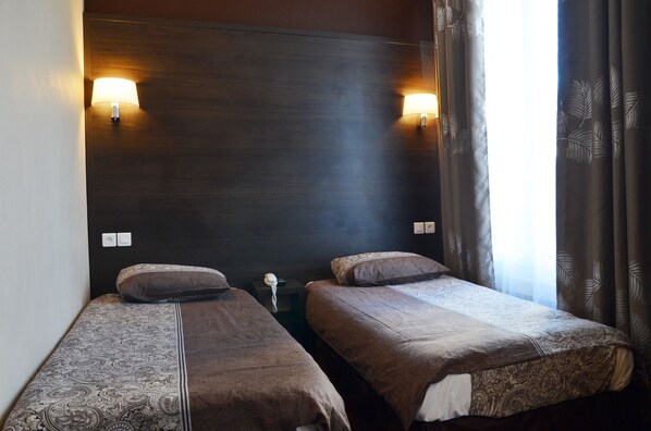 A hotel room with two single beds, a dark headboard, wall-mounted lamps, a bedside table with a lamp, and a window with curtains. - Hôtel du Parc Even (Malakoff)