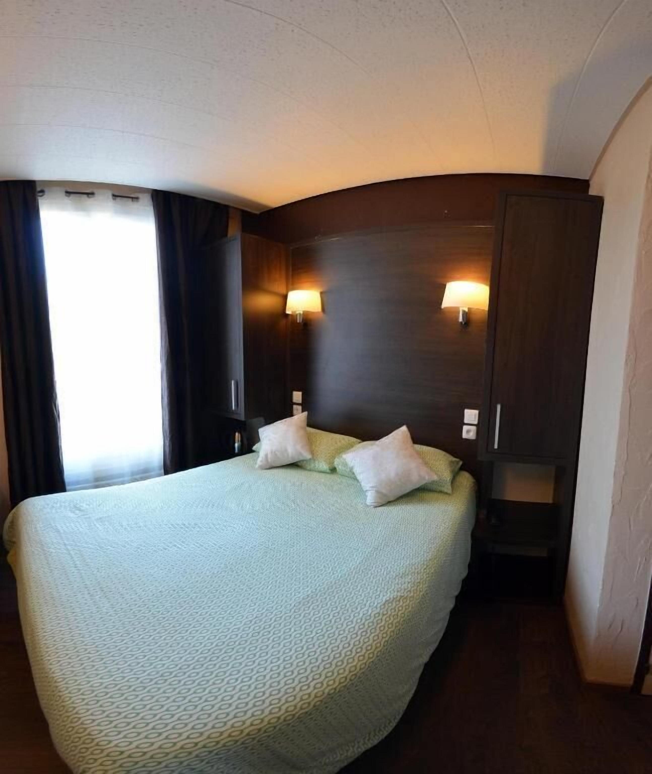 A hotel room with a large bed, two bedside lamps, dark wooden headboard, and a window with curtains.