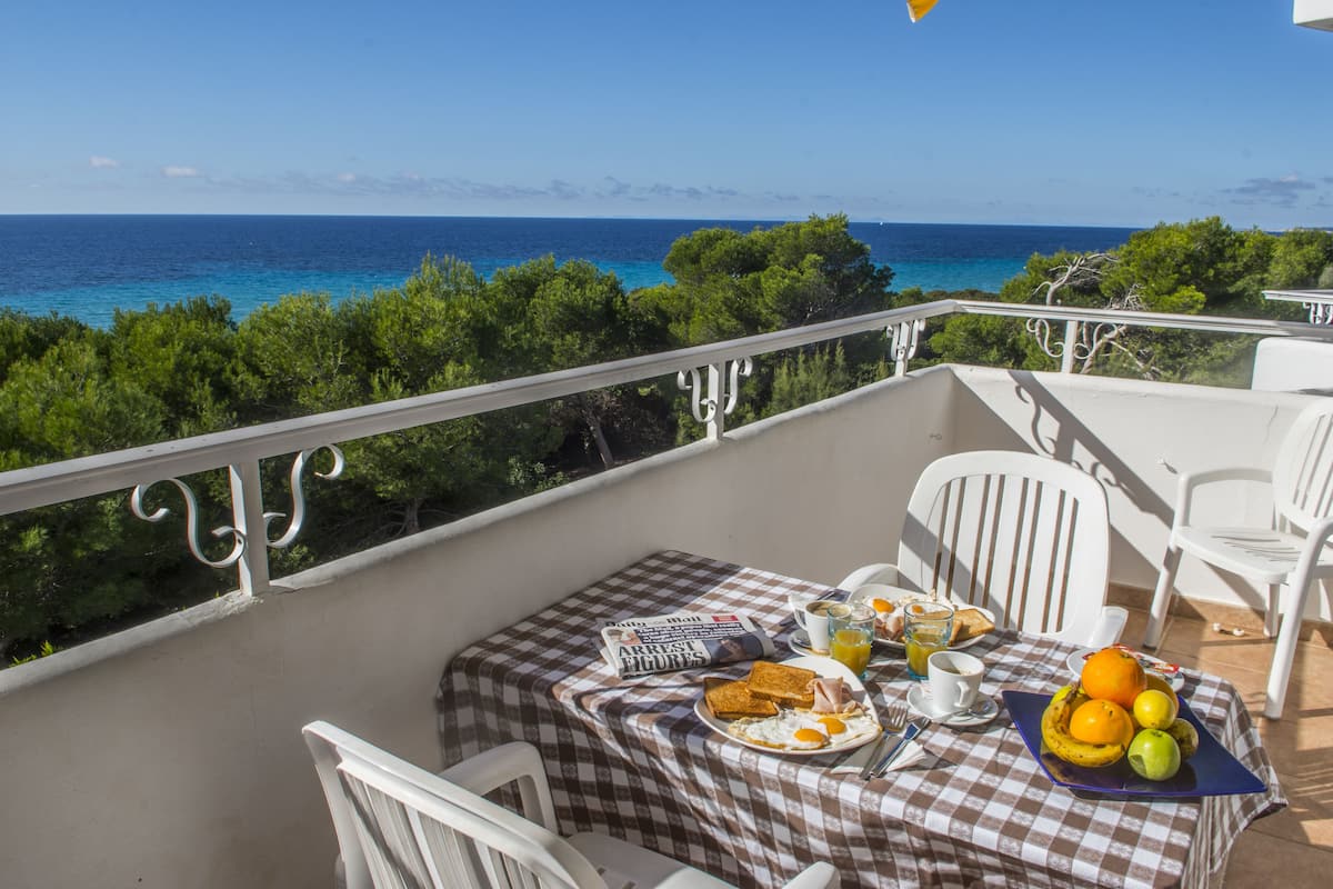 Ein Balkon mit karierter Tischdecke, weißen Stühlen und einer Frühstücksecke mit Toast, Eiern, Obst und einer Zeitung.