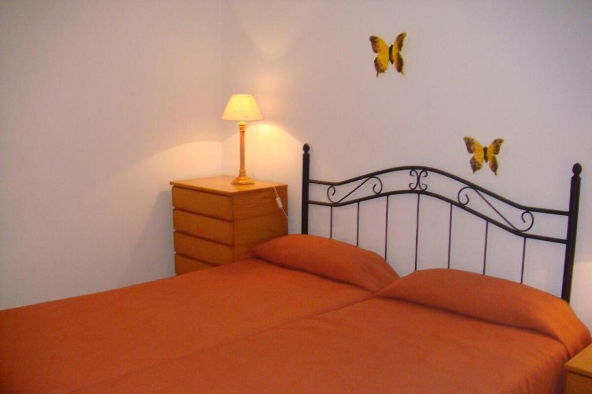 Un lit recouvert d’une couvre-lit orange, une table de chevet en bois avec une lampe, et deux décorations murales en forme de papillons.
