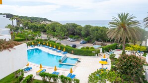A pool area with sun loungers and umbrellas, surrounded by greenery and a view of the sea. - Hamilton Court  Apartamentos (Es Migjorn Gran)