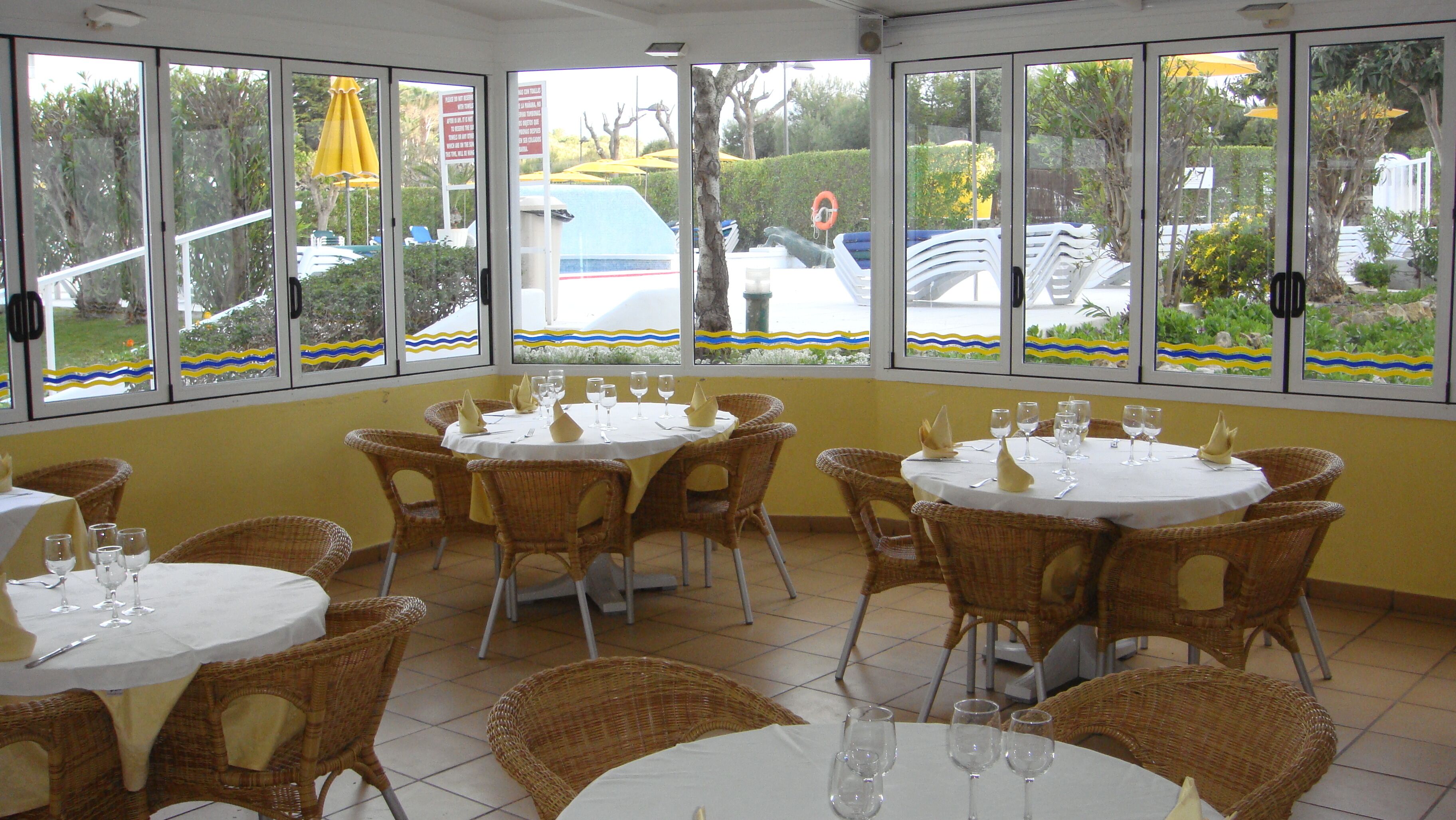 A restaurant with multiple tables and wicker chairs, large windows with decorative borders, and a view of the outside.