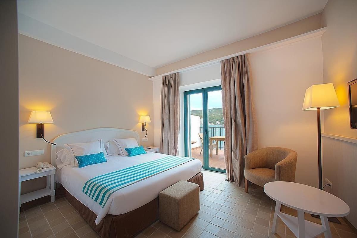 A hotel room with a bed, bedside lamps, a chair, a table, and a view of the outdoors.