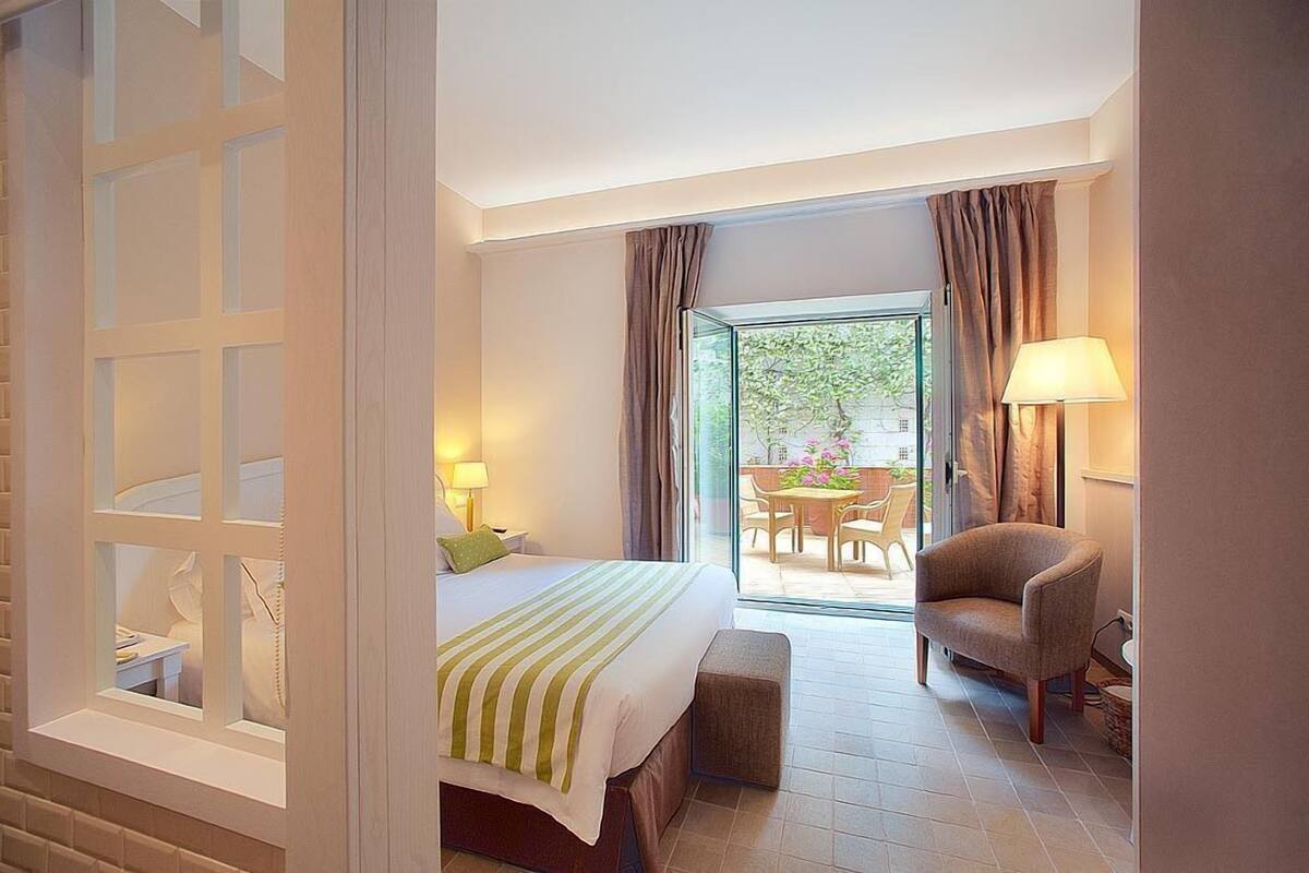 A hotel room with a bed, a chair, a table, and a view of an outdoor area with a table and chairs.