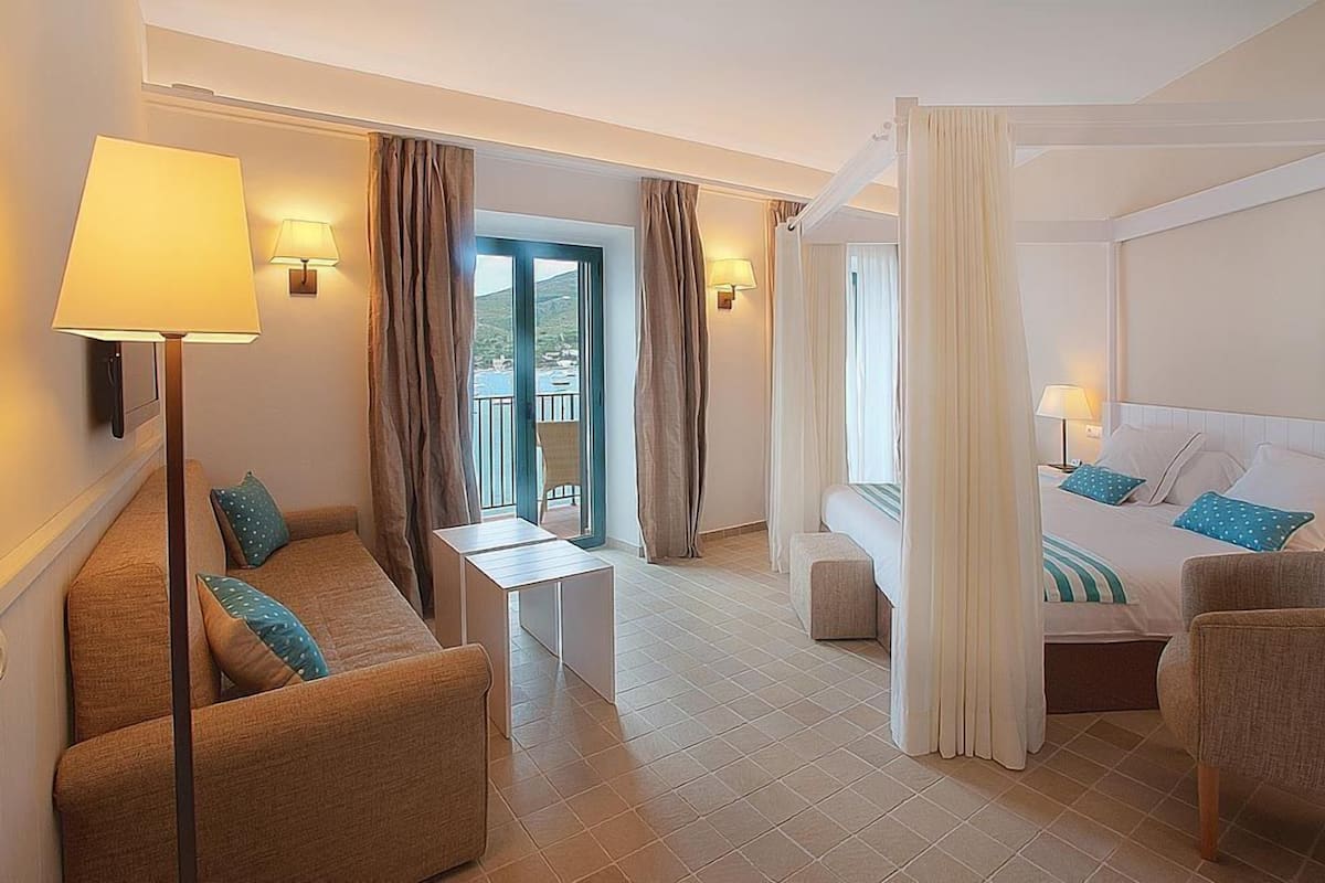 A hotel room with a sofa, a bed, a small table, and a view of the sea through a window.