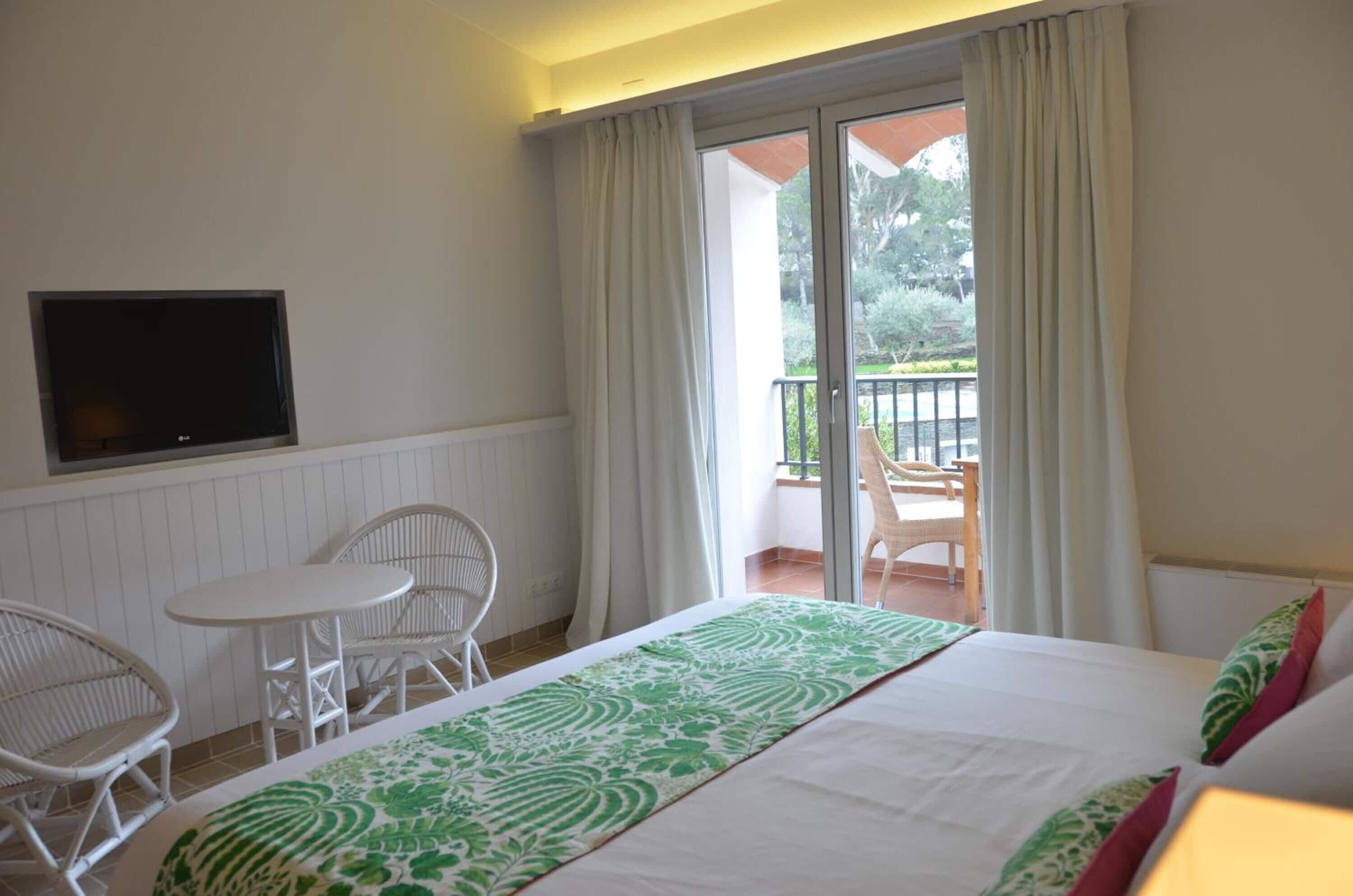 Habitación de hotel con cama, televisor, balcón con sillas y vista a zonas verdes.