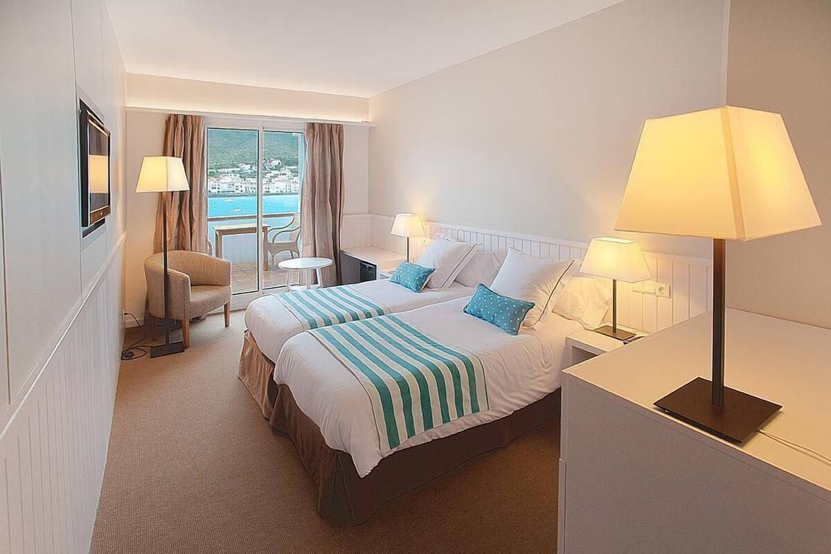A hotel room with two beds, a desk, a chair, a small table, and a view of the sea.