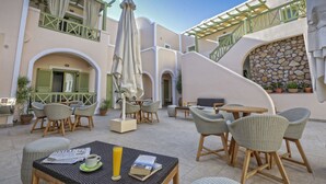 An outdoor seating area with wicker furniture, a table with a newspaper and a glass of orange juice, and a staircase with a stone wall. - Hotel Anassa (Santorini)