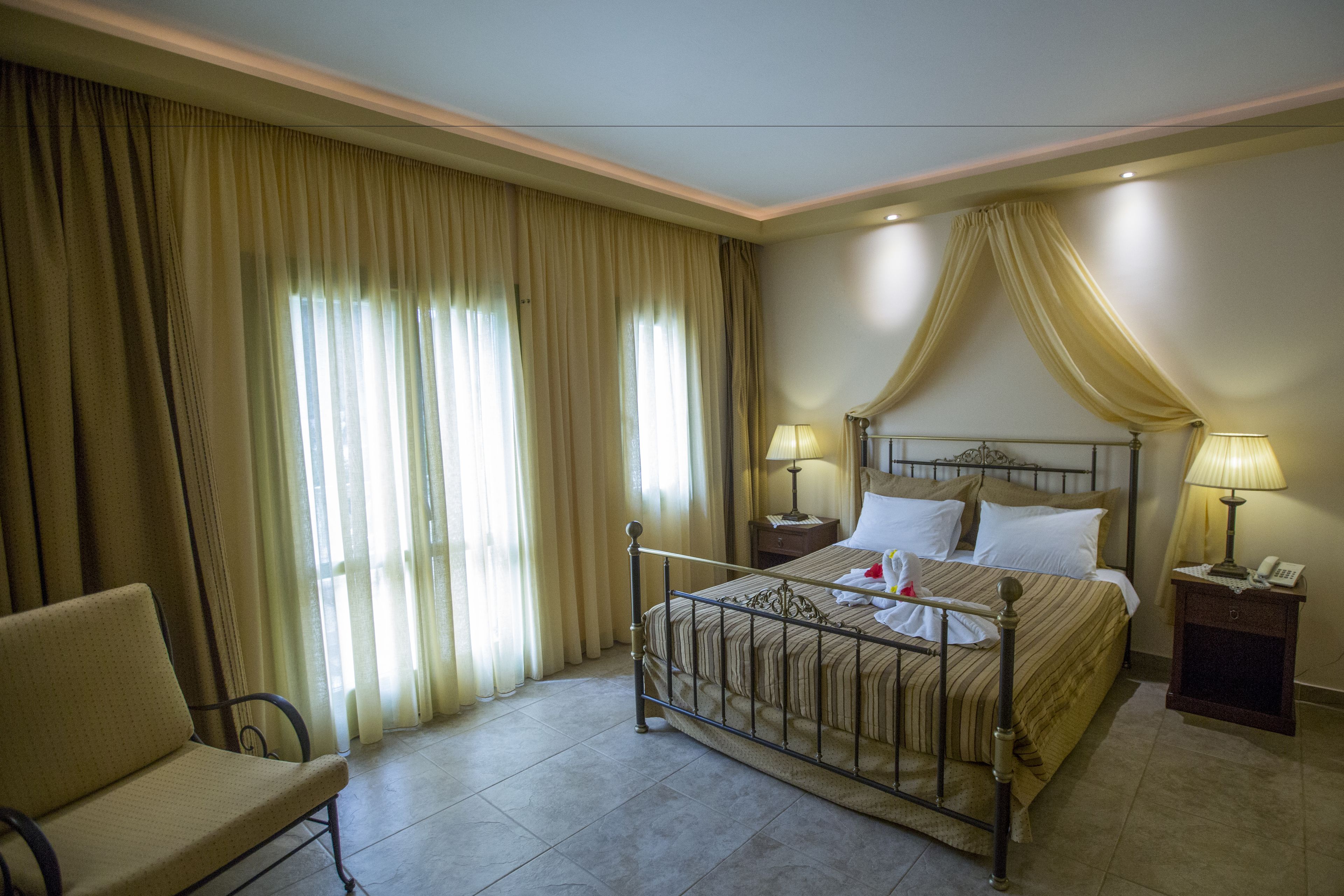 A hotel room with a bed, two bedside tables with lamps, a chair, and large curtains.