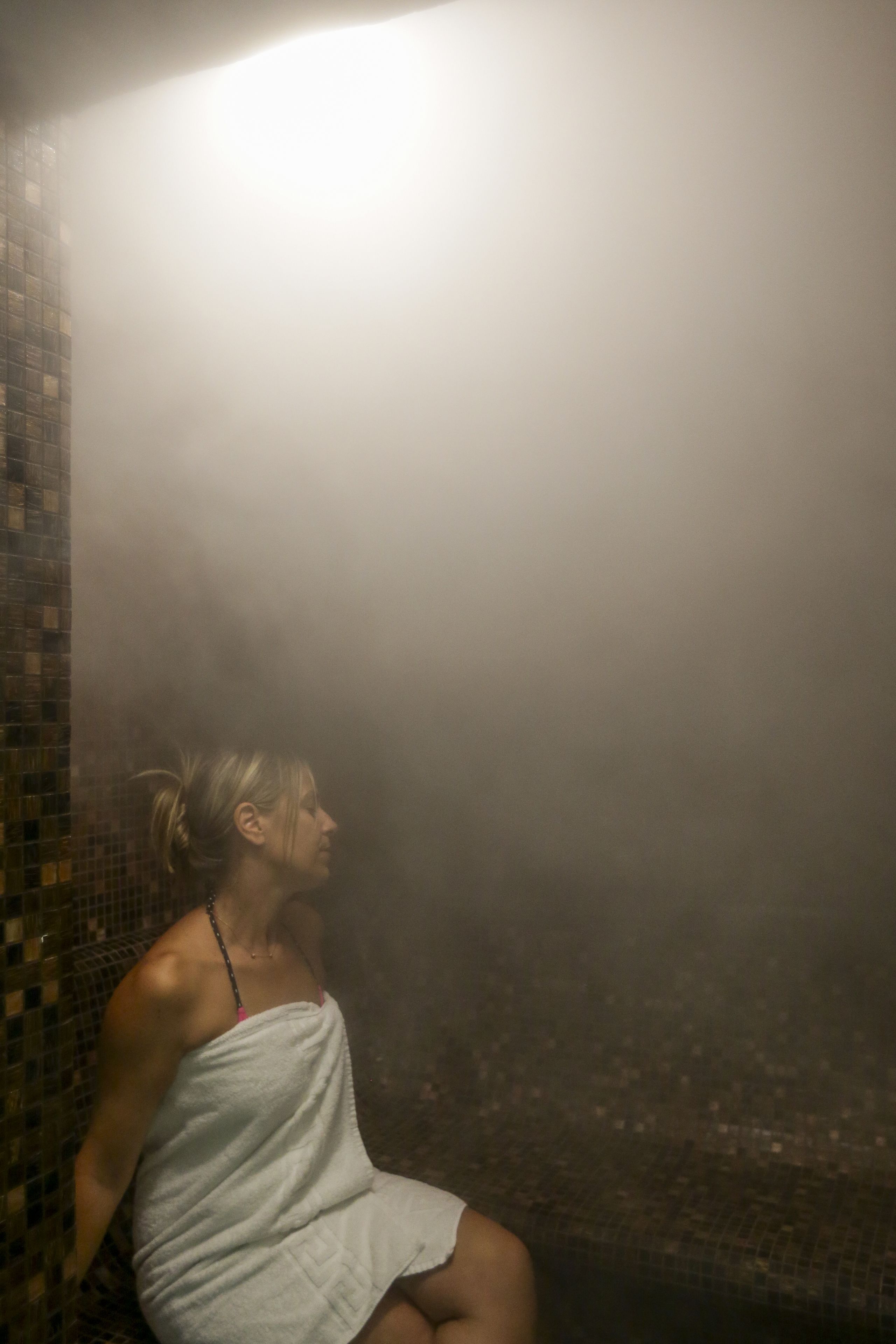 A person sitting in a steam room with a tiled wall and a light source above.