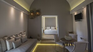 A modern hotel room with a sofa, a dining table with chairs, a television, and a large window. - Hotel Anassa (Santorini)