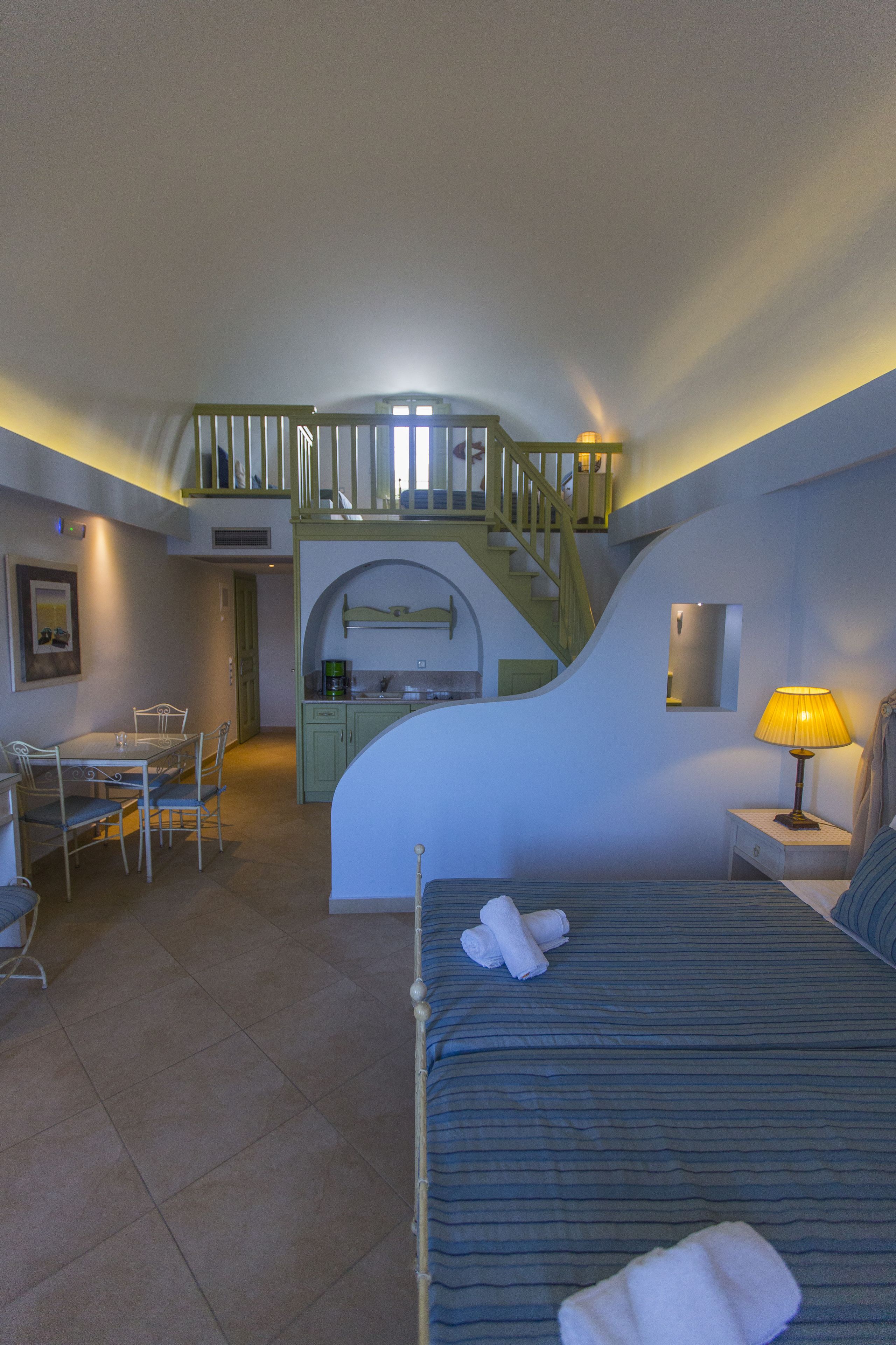 Ein kompaktes Hotelzimmer mit einem Bett, einem Schreibtisch, einem Stuhl, einer kleinen Küchenzeile und einer Treppe zu einer erhöhten Ebene.