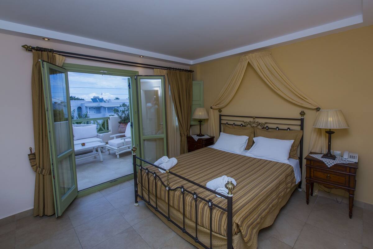 A hotel room with a bed, a nightstand, a lamp, and a view of the outside.