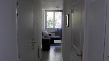 Un couloir étroit, avec un canapé de couleur sombre et une petite table, qui mène à une pièce dotée d’une grande fenêtre et d’un tableau encadré accroché au mur.