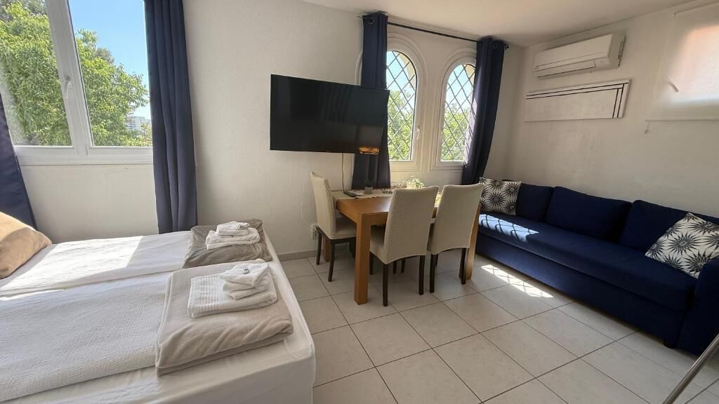 A room with a bed, a sofa, a dining table with chairs, a television, and windows with curtains.