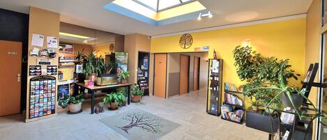A hotel lobby with a skylight, a reception desk, potted plants, a bulletin board, and a wall with a tree logo.