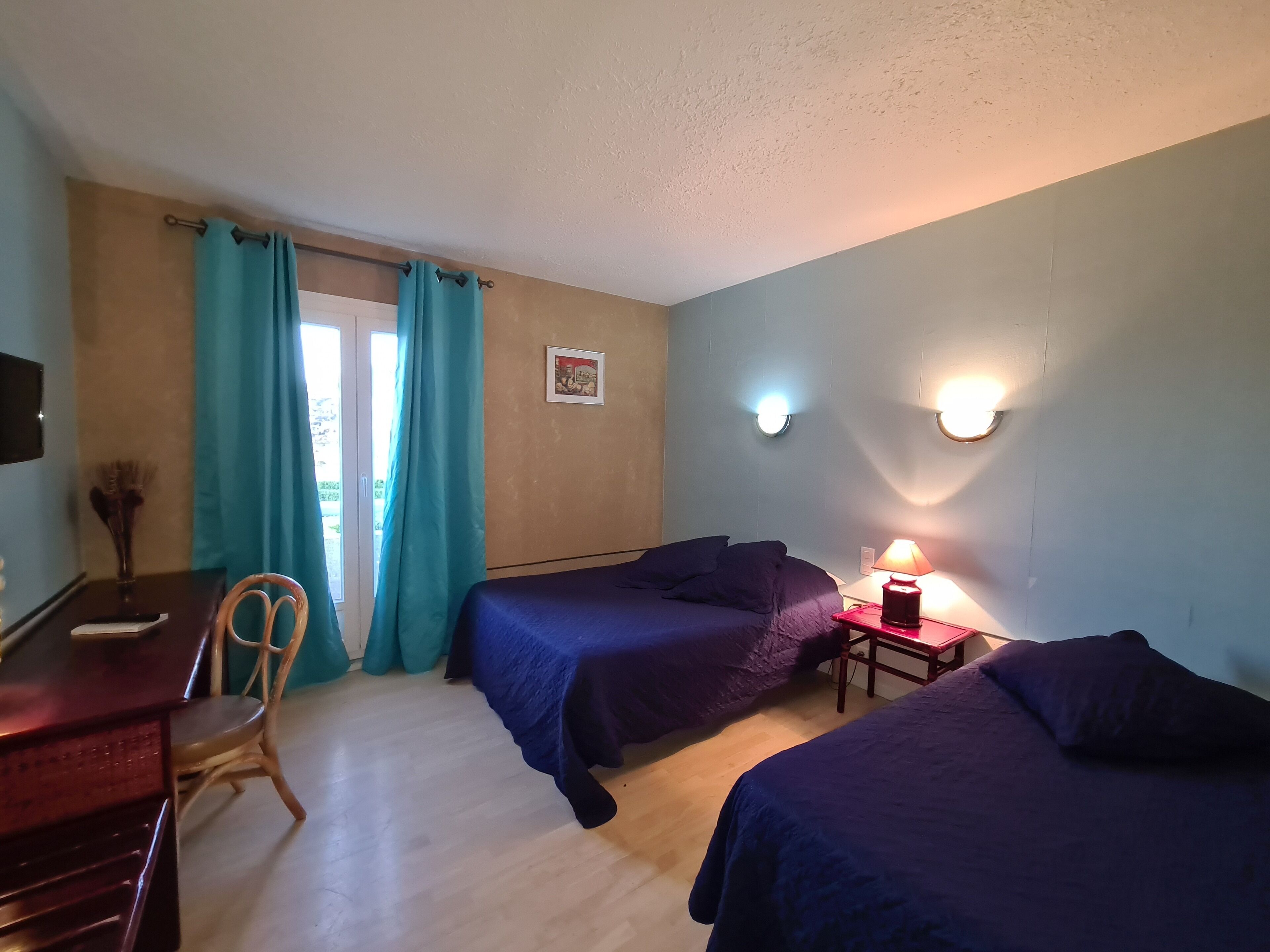 A room with two beds, a desk, a chair, a table with a lamp, and a picture on the wall.