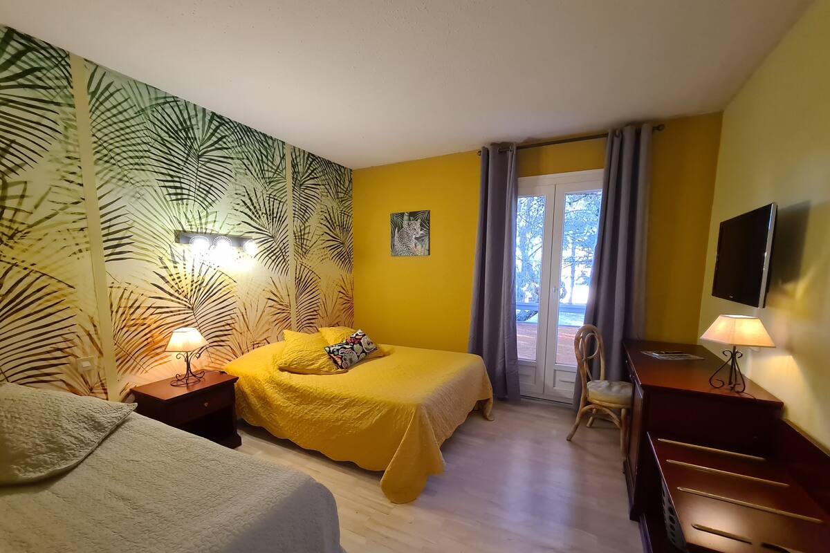A hotel room with two beds, a desk, a TV, and a balcony with a view of trees.