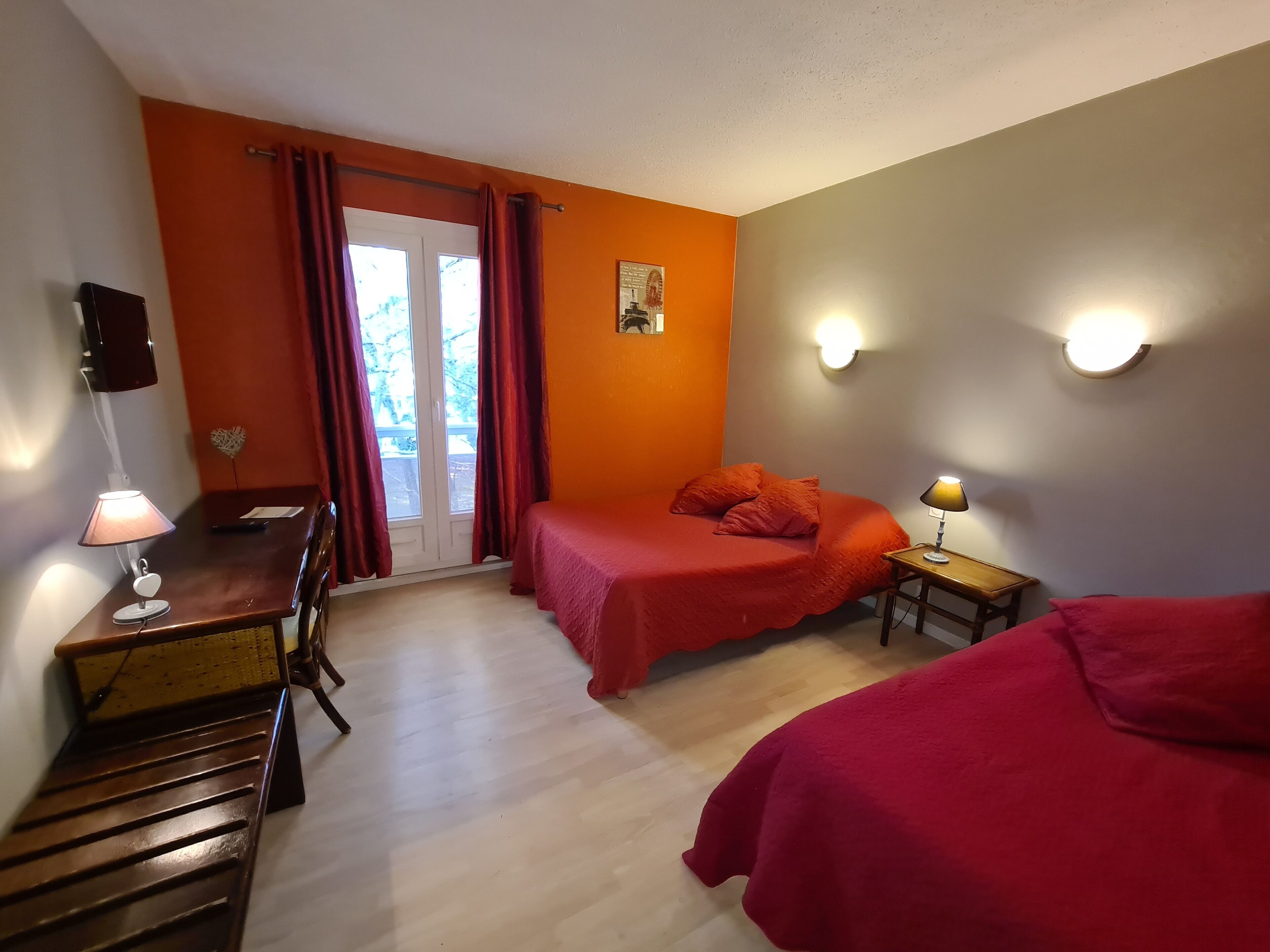 A room with two beds, a desk, and a chair. There are two wall-mounted lamps and a window with curtains.